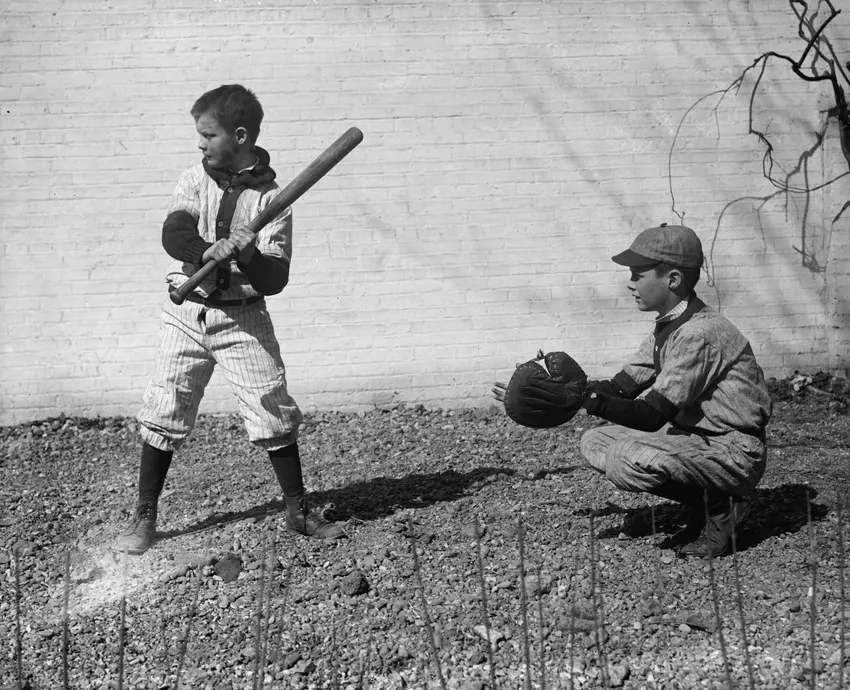 Two boys in old wool uniforms, one batting and one catching, against a brick wall
