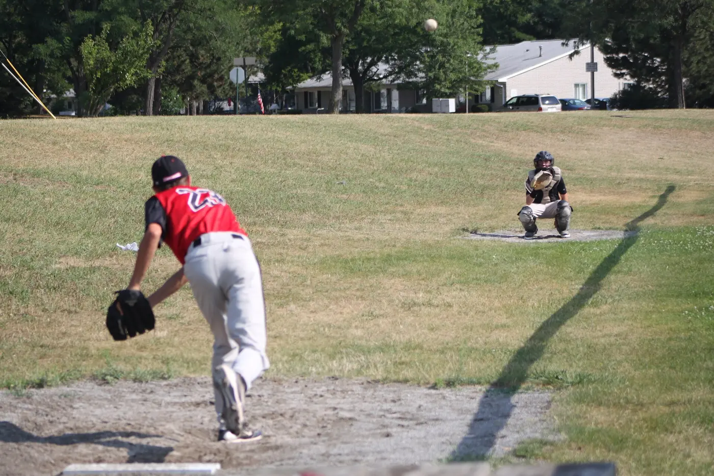 A youth pitcher in a red jersey throwing to a catcher on a grass field